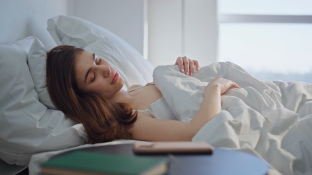 Žena leži u krevetu, dok je telefon pored nje na stolu