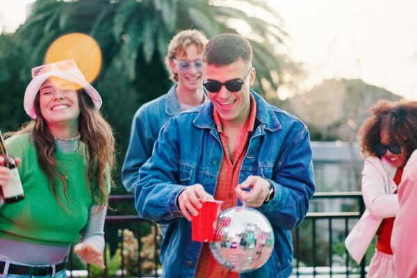 Ideje za vikend sa društvom. Grupa mladih ljudi uživa u vikend druženju na terasi, smeju se, piju piće i plešu uz muziku