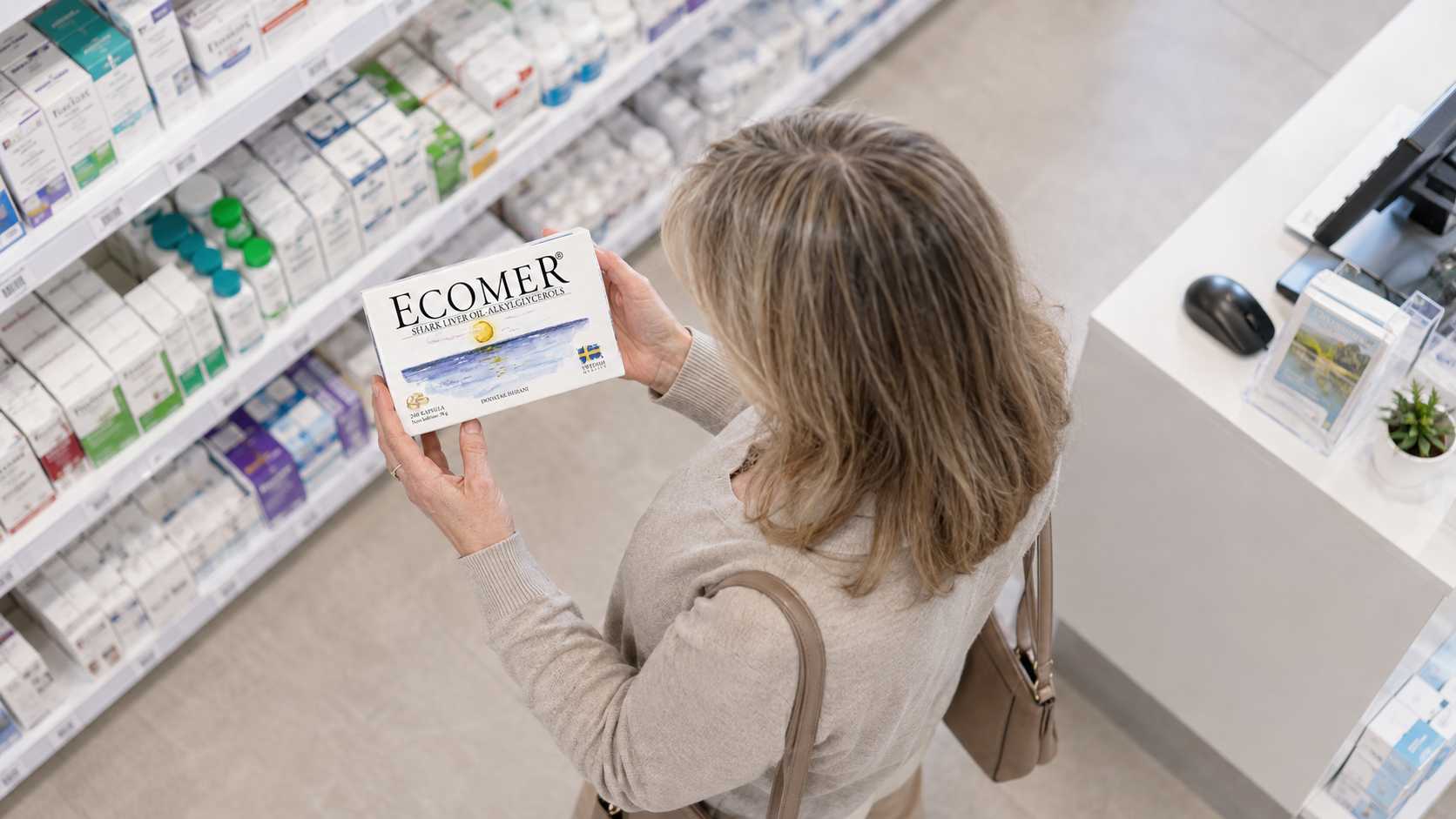 Žena u apoteci iz ptičje perspektive čita deklaraciju na kutiji Ecomera.