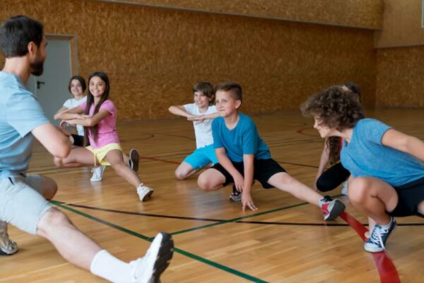 Nastavnik fizičkog vaspitanja i grupa dece školskog uzrasta rade vežbe istezanja nogu u sportskoj sali sa drvenim podom