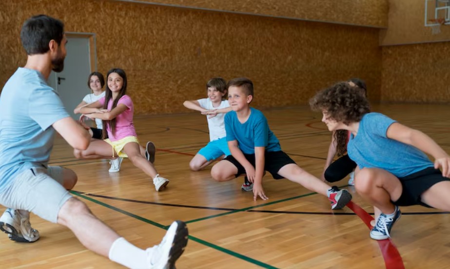 Nastavnik fizičkog vaspitanja i grupa dece školskog uzrasta rade vežbe istezanja nogu u sportskoj sali sa drvenim podom