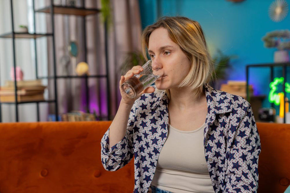 Žena kratke plave kose sedi na narandžastoj sofi i pije vodu iz staklene čaše